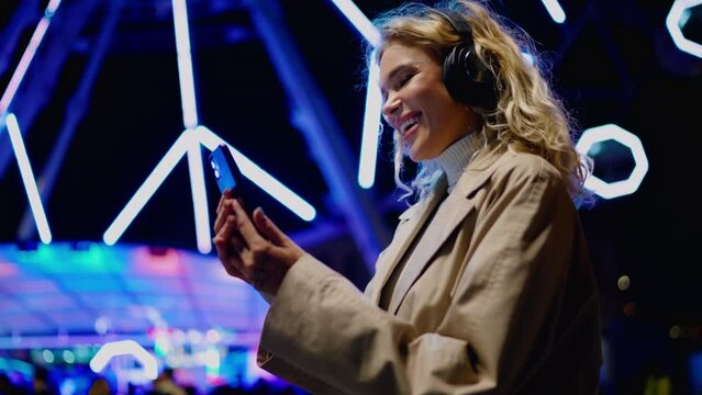 Attractive millennial female in headphones enjoying music playlist while standing against backdrop of colorful illuminating ferris wheel. Young woman blogger uses smartphone app for social media chat