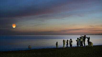 Silhouettes of people lighting Chinese lanterns on the beach, at dusk.