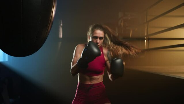 Portrait of fit, caucasian lady in her 20s warming up before the training. Close-up view of young and confident girl practicing her kick in boxing gloves. High quality 4k footage