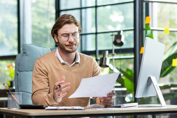 A serious young man, a businessman, a programmer sits in the office at a computer. Works with...
