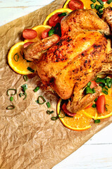 Grilled whole chicken on oranges and tomatoes. View from top. White wooden boards and baking paper. Vertical photo.
