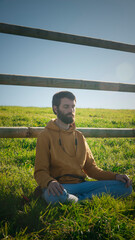 Hombre barbudo meditando junto a valla de madera en pradera de hierba