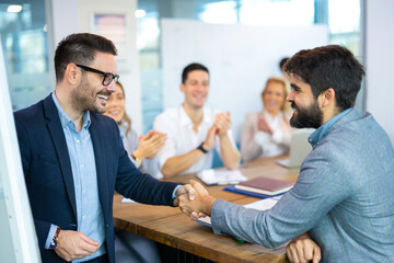 Business men shaking hands while co-workers in the background applauding and celebrating successful...