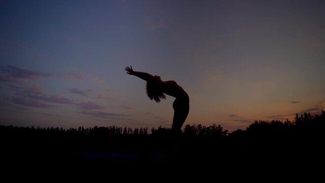 Woman doing yoga stretching, bending against sunset sky, backlit silhouette
