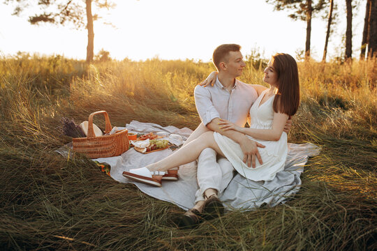 A couple in love, husband and wife, are sitting on a picnic in a field under trees. Lovestory at sunset in summer. Warm family shooting in nature