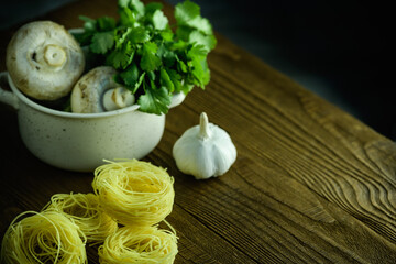 Raw pasta nests, mushrooms in bowl and garlic on wooden table on dark background