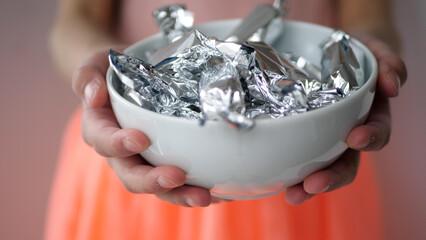 Close-up shot of a girl's hands holding a white ceramic bowl full of candy in silver wrapping paper - 572793430