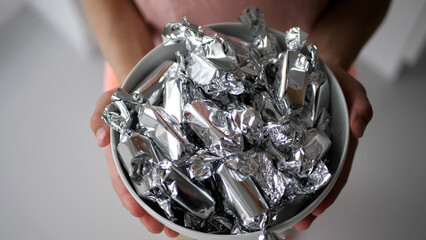 Close-up shot of a girl's hands holding a white ceramic bowl full of candy in silver wrapping paper - 572793466