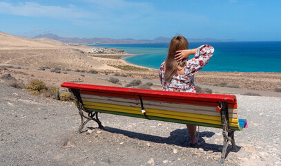 Mujer joven con el pelo largo y rubio y con un vestido sentado en un banco con colores lgtbi en la espalda frente a la playa de arena blanca y aguas turquesas de Sotavento Fuerteventura Islas Canarias