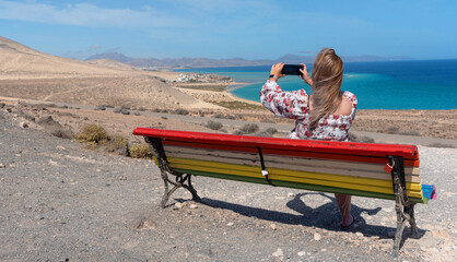 Mujer joven con cabello largo y rubio y vestido tomando una foto en un banco con colores lgtbi frente a la playa de arena blanca y aguas turquesas de Sotavento Fuerteventura Islas Canarias