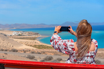 Mujer joven con cabello largo y rubio y vestido tomando una foto en un banco frente a la playa de arena blanca y aguas turquesas de Sotavento Fuerteventura Islas Canarias en un día soleado
