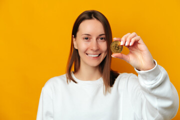 Smiling young woman is showing to the camera a golden coin of bitcoin.