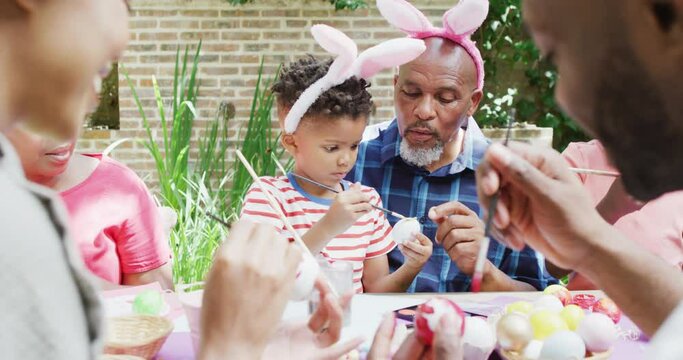 African american grandfather and grandson painting easter eggs with family in garden, slow motion