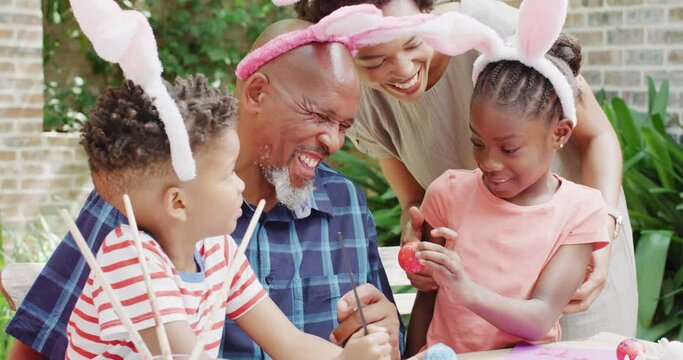 Happy african american brother, sister and mother painting easter eggs with grandfather, slow motion