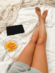 woman in cozy short pants barefoot lying on bed next to her digital Tablet, in ear headphones, bowl of fresh mango and granola