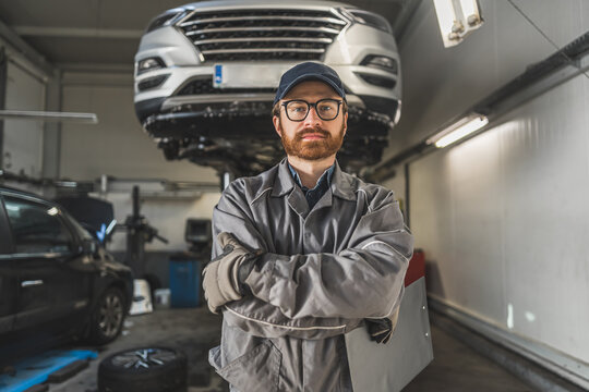 Repair shop concept. A mechanic in unnoform standing in the center and posing for a picture with his arms crossed. Car on a lift in the background. High quality photo