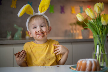A cute little boy wearing bunny ears on Easter day is eating a chocolate Easter bunny. A child...