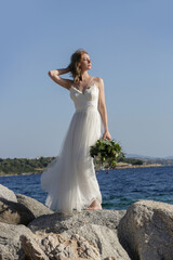 Fototapeta na wymiar Bride in long white wedding dress standing on windy sea rock shore holding lush green wedding bouquet with boho twist