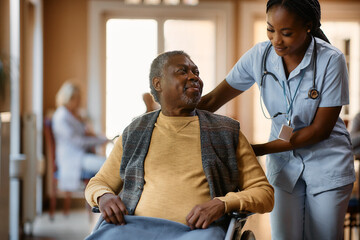 Young black nurse assists senior man in wheelchair at nursing home.