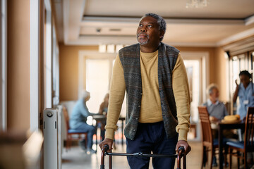 Pensive black senior man with walker looks through window at residential care home.