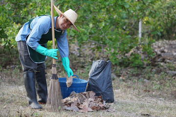 Asian male  gardener holds broom , basket, black garbage bag to clean dry leaves in garden.        ...