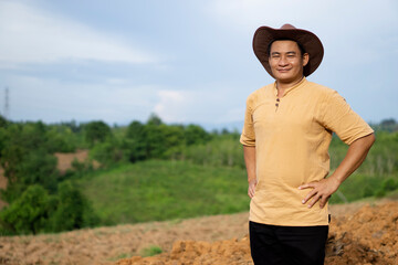 Portrait of Asian man farmer wears hat, brown shirt, puts hands on hips, feels confident, stands at agriculture land  before growing crops. Concept, agriculture occupation. Thai farmer. Copy space.
