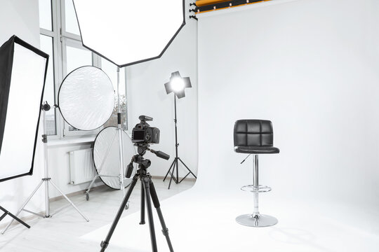 Tripod with camera, bar stool and professional lighting equipment in modern photo studio