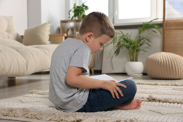 Boy with poor posture reading book on beige carpet in living room. Symptom of scoliosis