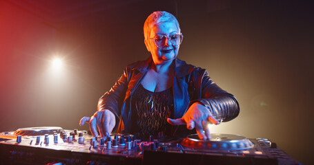 A cool caucasian dj grandma is composing a mix at turntables controller. Authentic mature woman...