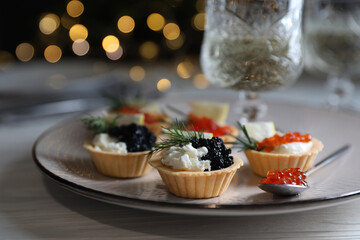 Delicious tartlets with red and black caviar served on white wooden table against blurred festive lights, closeup. Space for text