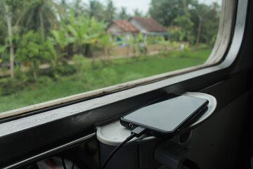 cell phone that is being charged during the trip available on the train with a window background