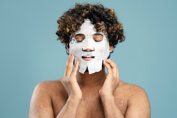 Headsot, Curly-haired ethnic man with tissue facial mask on blue background. Skin care concept