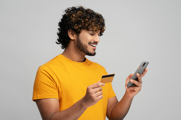 Smiling Asian ethnic man holding credit card and using phone, transferring money via mobile banking