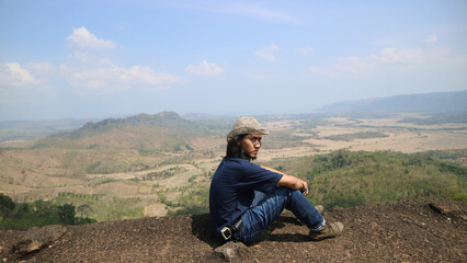 Naklejka na ściany i meble A man sitting on a huge rock on the top of hill, enjoying the views. Explorer scenes.