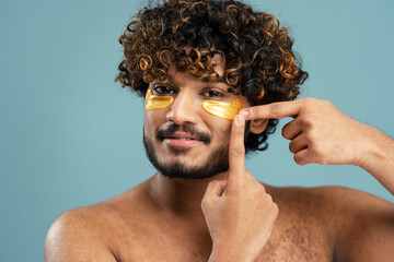 Young Indian man face applying hydrating undereye patches, morning routine concept on blue backdrop