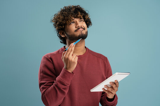 Pensive Indian man dreamily looking up posing with notepad in his hand, isolated on blue background
