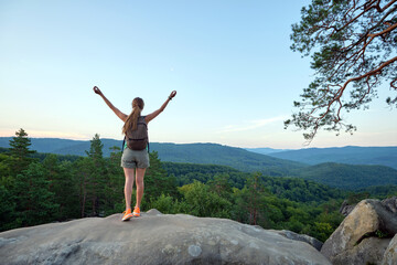 Sportive woman standing alone on hillside trail with raised up arms. Female hiker enjoying view of evening nature from rocky cliff on wilderness path. Active lifestyle concept