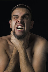 Portrait of a bearded man with open mouth, strangling himself with both hands. Black background. The man grimaces and looks into the camera. Shirtless man. Concept of people and emotions. Close up.