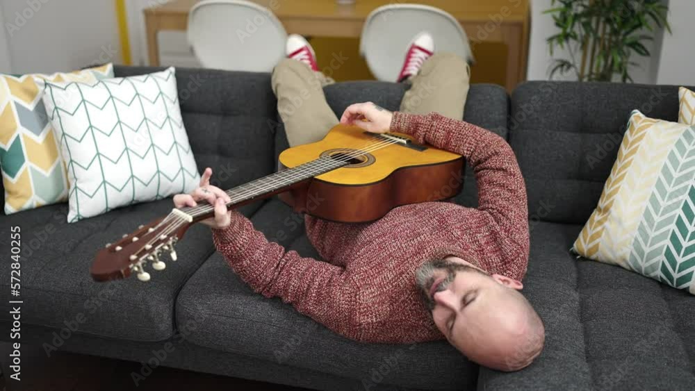 Poster young bald man playing classical guitar lying on sofa at home