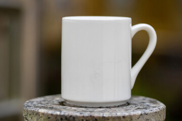 White mug, mock up, empty space for artwork, text, standing on stone, outdoors, green grass, trees, sky in the background, soft sunlight, nature