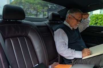 Confident senior business man in suit wearing eye glasses sitting on car backseat and writing business plan in the book during go to office. Elderly businessman executive working on the road.