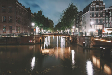 Naklejka premium Amsterdam canal, bridge, and medieval houses in the evening, Bright cityscape of Amsterdam with night lights reflection in the water, Night landscape with houses and canal in amsterdam