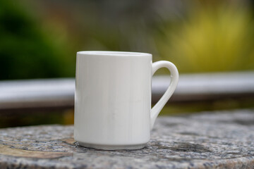 White mug, mock up, empty space for artwork, text, standing on stone, outdoors, green grass, trees, sky in the background, soft sunlight, nature