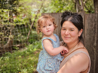 Fototapeta premium Mom holding daughter and looking at camera in natural backyard