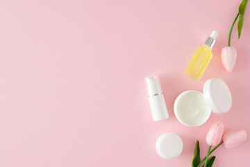 Skincare beauty concept. Flat lay photo of white cosmetic tubes, cream jars, serum bottles and spring flowers on pastel pink background with copyspace. Skin care products mockup.