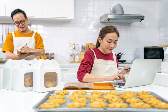 Asian man and woman bakery shop owner working on laptop computer and listing customer order in kitchen. Bakery chef making cookies for selling online delivery. Small business food and drink concept.