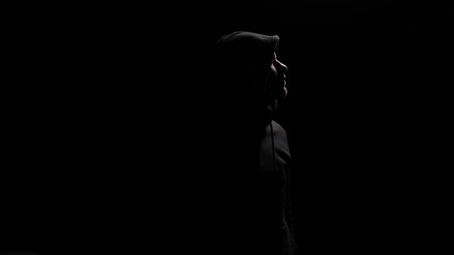 Silhouette of mysterious man in black hoodie looks up and then at camera, with face hidden. conveying mystery, drama, intrigue, psychological state. Isolated portrait of a stranger or anonymous man.