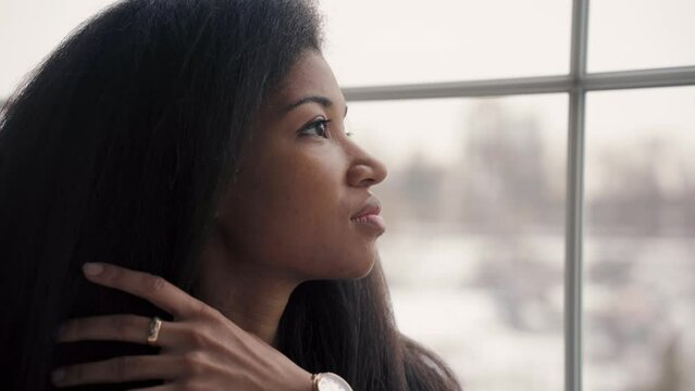 An African girl stands near the window and straightens her hair.