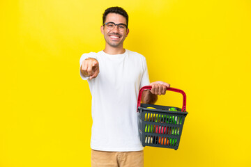 Young handsome man holding a shopping basket full of food over isolated yellow background surprised and pointing front