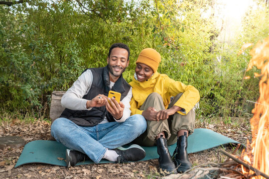 Young multiethnic couple sitting around the campfire looking at mobile phone. High quality photo